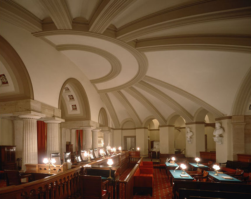 Above, a vaulted, semi-circular ceiling divided into ten ribbed sections. Below 
					and to the left are several high-backed chairs behind a wooden railing. Furniture on the right includes four tables covered in green fabric. Photograph courtesy of the 
					U.S. Senate.