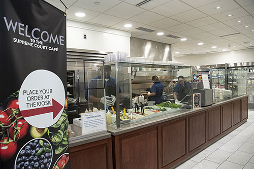 Sign displaying the words,“Welcome to the Supreme Court Café,” and “Place your order at the kiosk,” with an arrow pointing to the right. Café employees prepare food behind a counter.