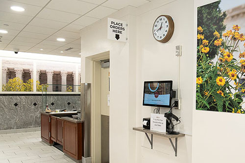"Place Orders Here” sign with a downward pointing arrow. Below and to the right of the sign is an electronic kiosk for placing orders and scanning grab-and-go items.