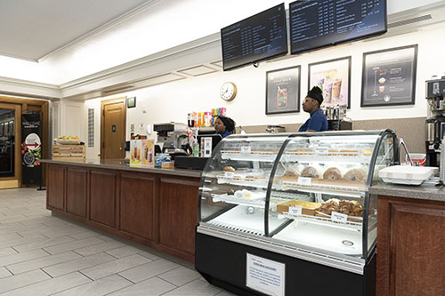Two employees stand behind the Starbucks counter. There is a menu hanging above and a selection of baked goods in a glass case.