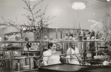 Elizabeth Mertens (left) and a fellow cafeteria 
                                                                                            employee behind a display featuring forsythia clippings and other spring foliage from Mertens's garden, circa 1950s.