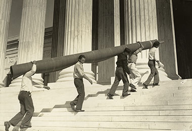 Five men carry a roughly 18-foot carpet roll on their shoulders up the front steps of the Supreme Court Building.